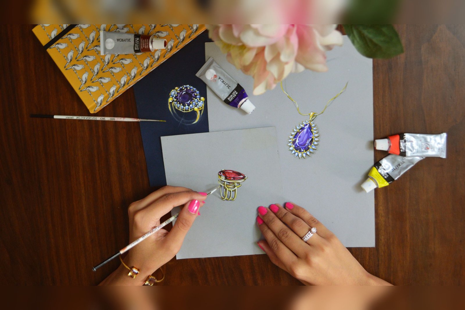 Hands painting gouache renderings of a ring and a pendant at a desk, beside paint tubes, a brush and a sketchbook.