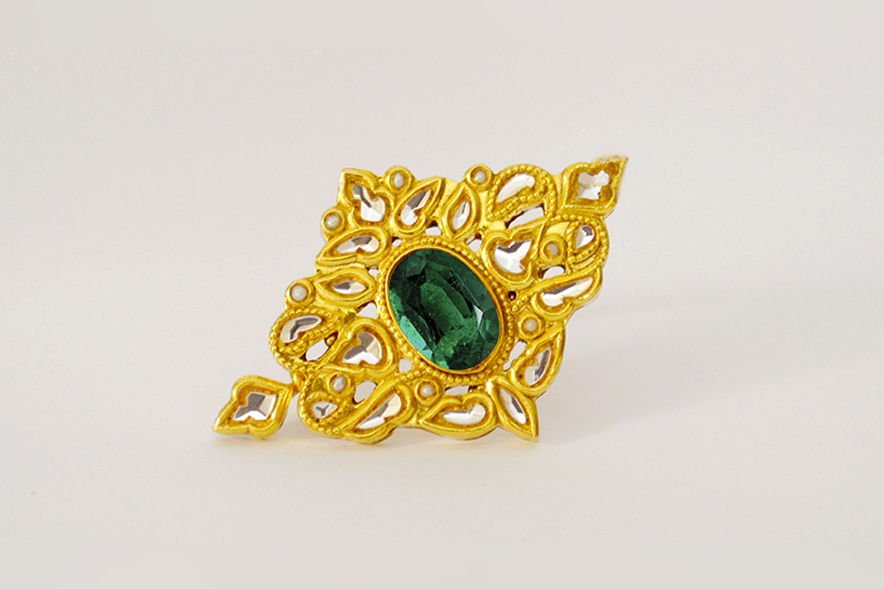 A Mumtaz kundan hair-clip set with a green emerald and framed by kundan petals, on an off-white ground.