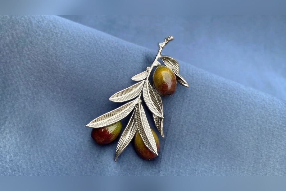 The Olive Branch brooch in silver with hand-painted olives, on blue cloth.