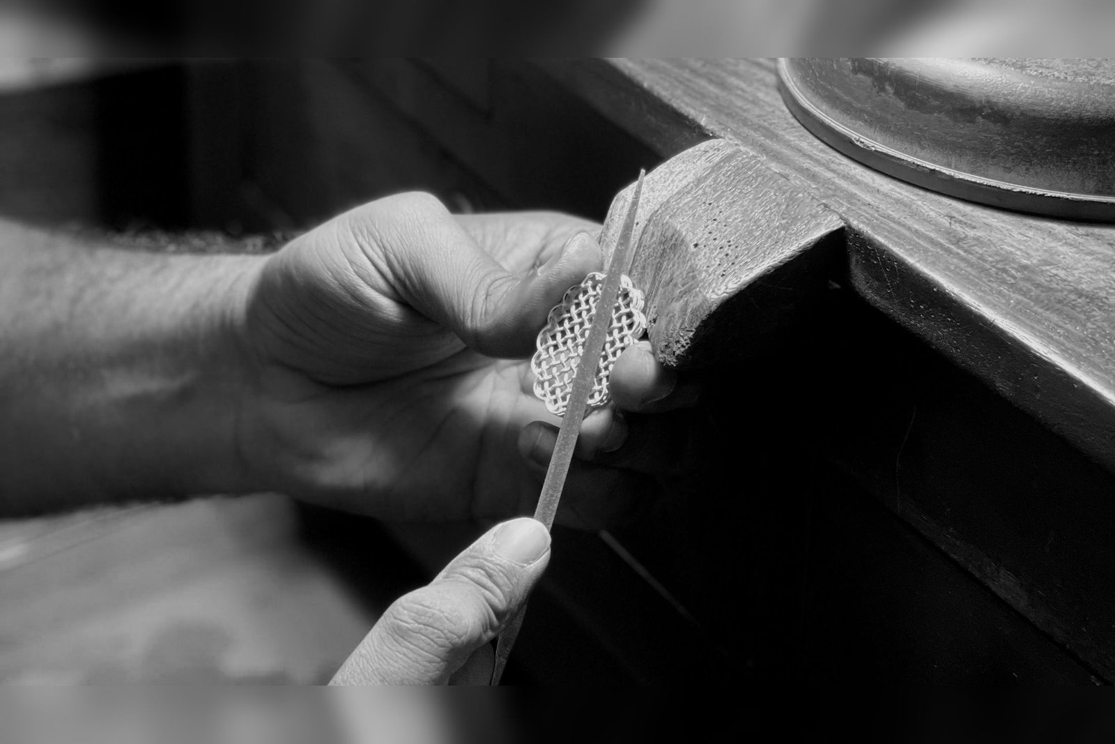 Black and white photograph of a hand filing the edge of a lattice filigree brooch against the wooden bench peg.