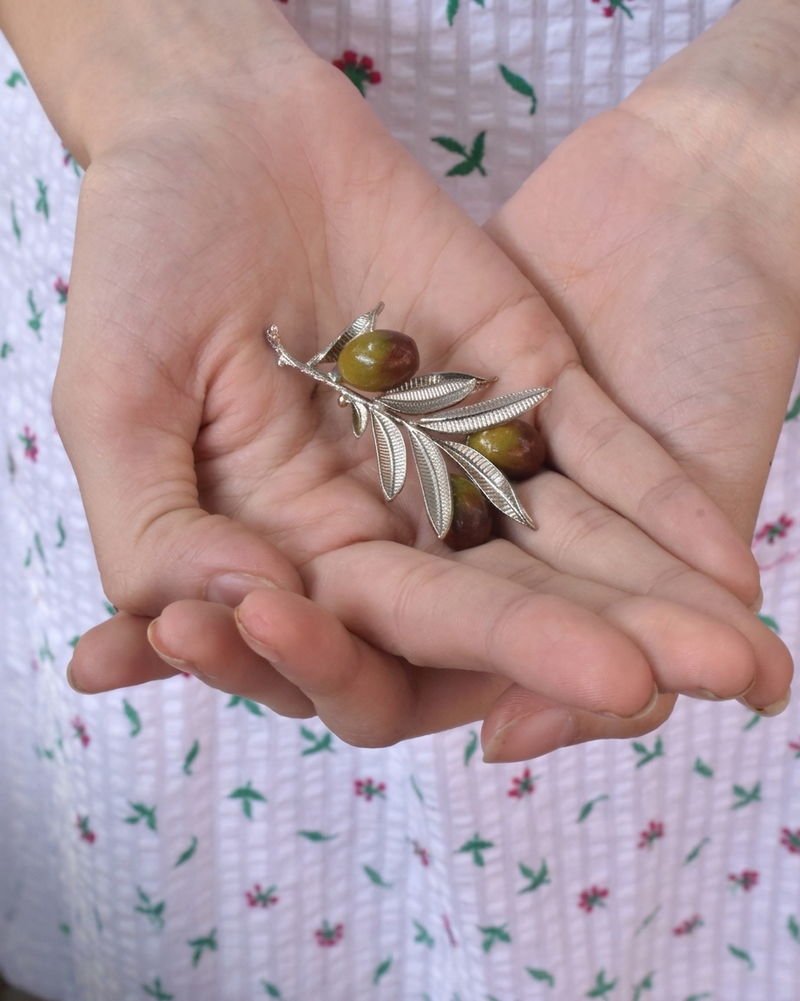 The Olive Branch brooch in silver with hand-painted olives, held in a cupped hand.