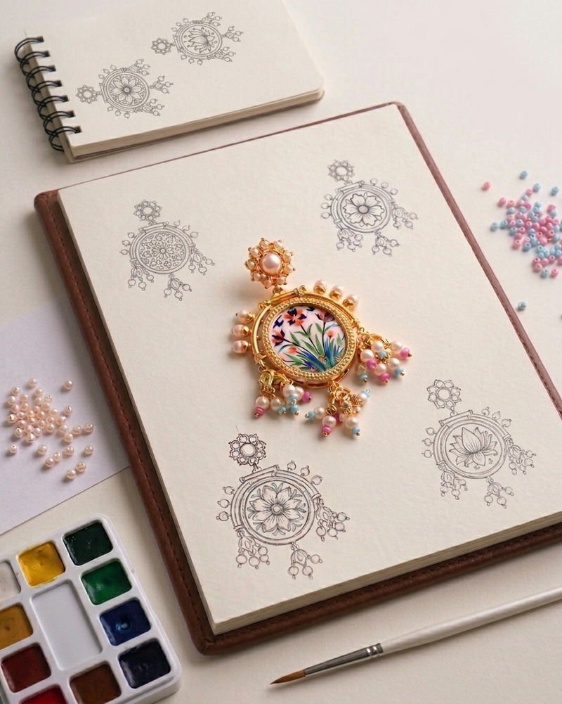 A finished Arabesque earring resting on an open sketchbook of pencil drawings, beside a watercolour palette, brush and loose pearls.