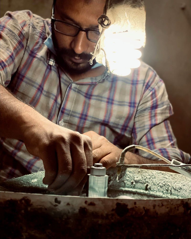 An artisan at the bench, working a piece of silver over a pitch bowl under a bare lamp.