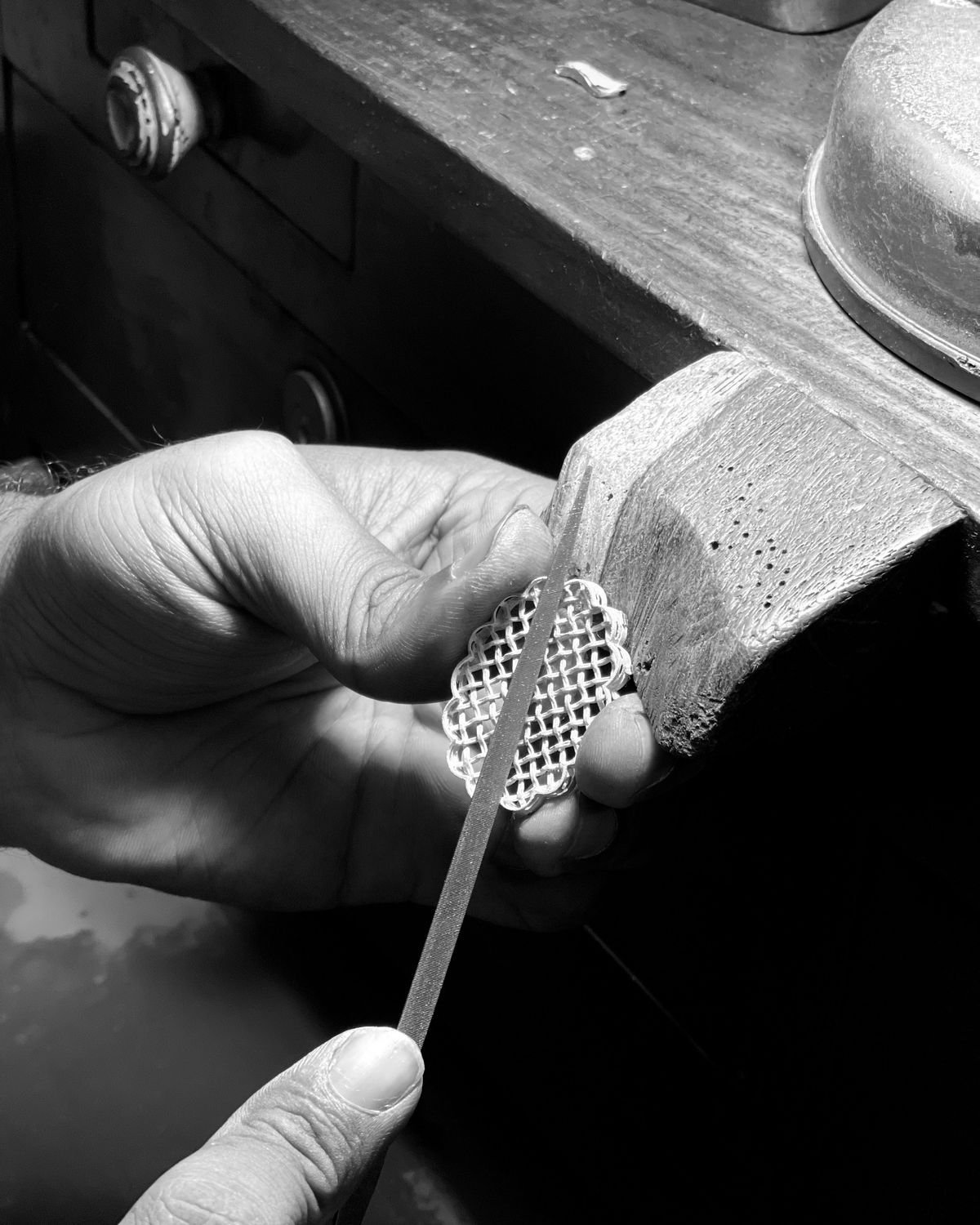 A hand holding a lattice filigree brooch against the wooden bench peg and filing its edge with a needle file.
