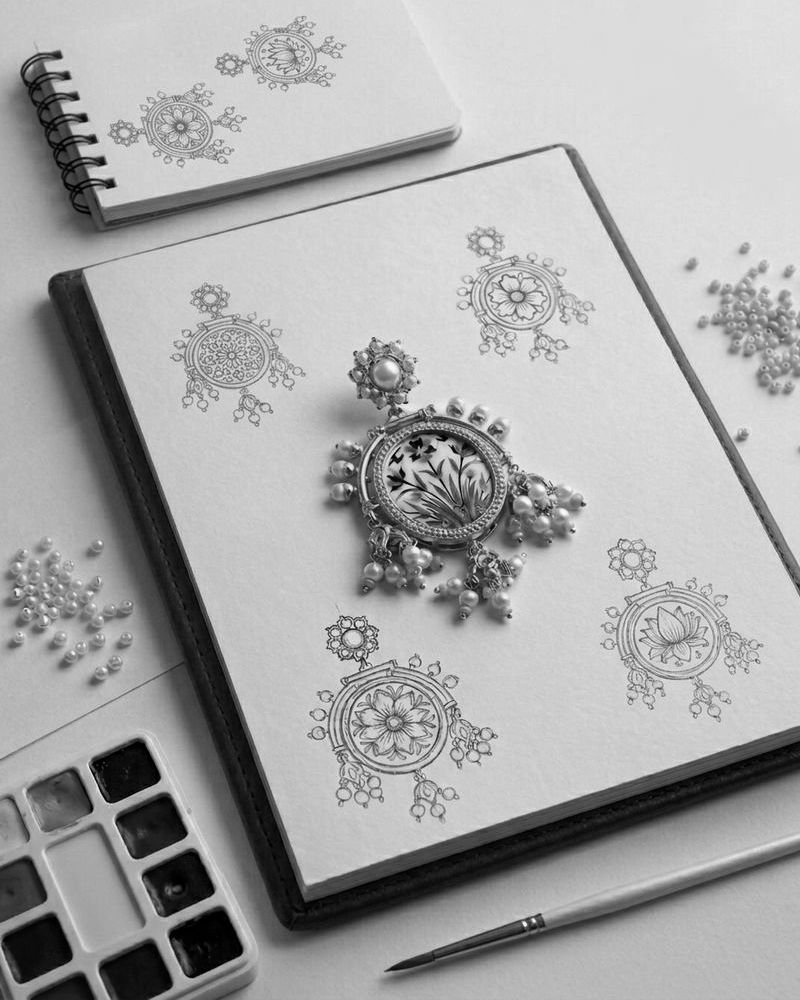A finished Arabesque piece resting on an open sketchbook of pencil drawings, beside a watercolour palette, brush and loose pearls.
